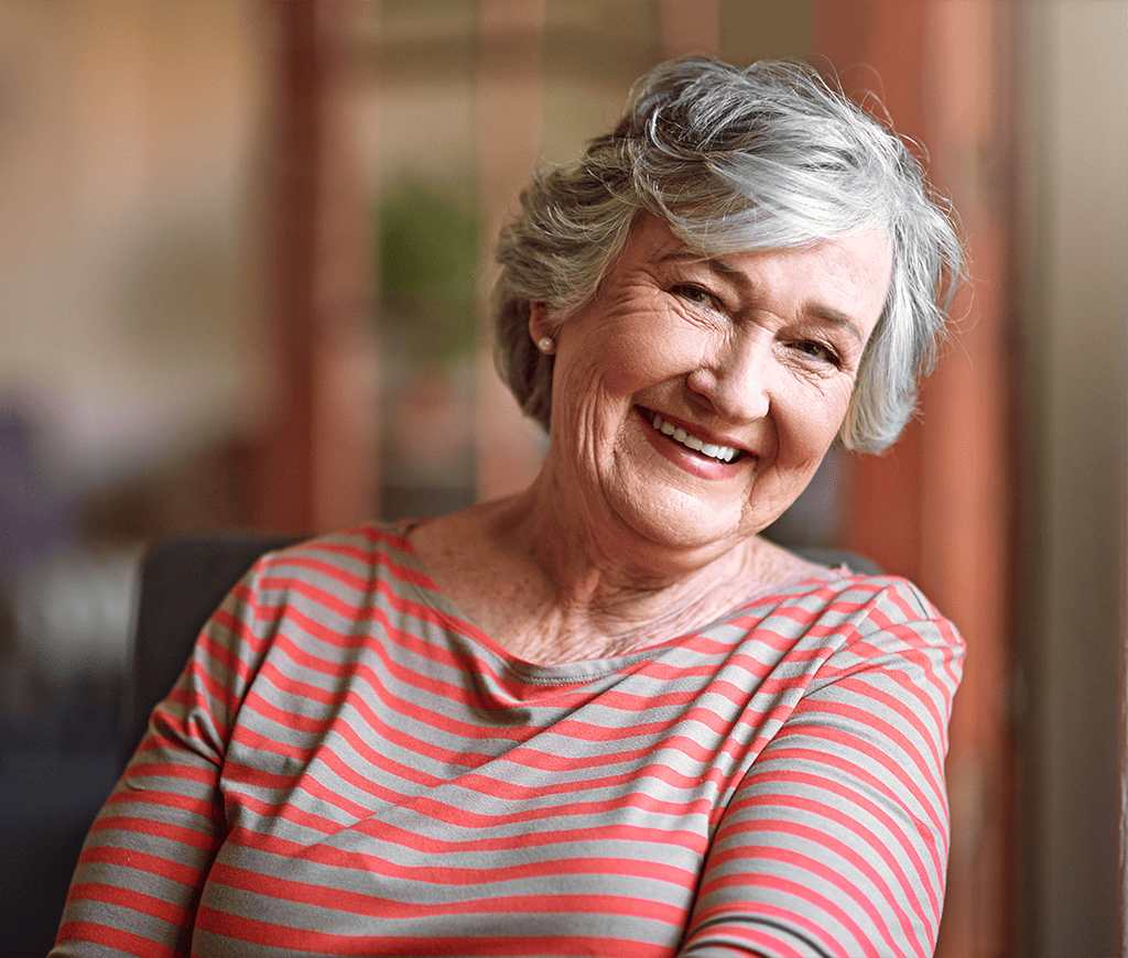 Elderly woman smiling in a striped shirt, seated indoors.